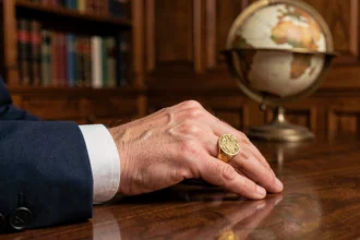 Chevalière homme en or gravée d'un blason familial portée sur la main gauche, posée sur un bureau en acajou dans un bureau classique
