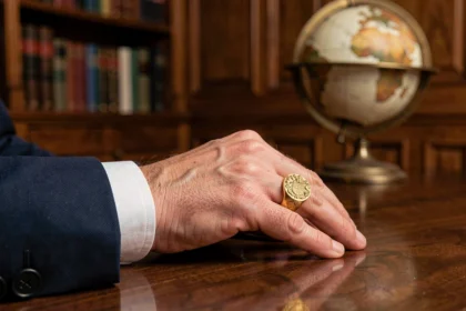 Chevalière homme en or gravée d'un blason familial portée sur la main gauche, posée sur un bureau en acajou dans un bureau classique