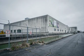 Vue extérieure d'une grande usine textile industrielle avec eaux usées verdâtres et végétation clairsemée sur asphalte fissuré